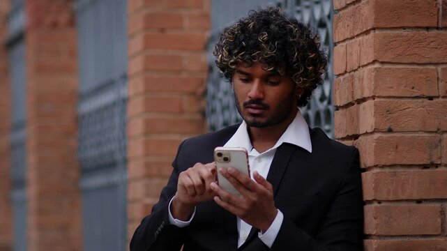 A man of Indian descent in a suit is lost in the city, looking for directions on his phone. He glances at the phone, then looks around to figure out the path he needs to take. Ideal for showcasing nav