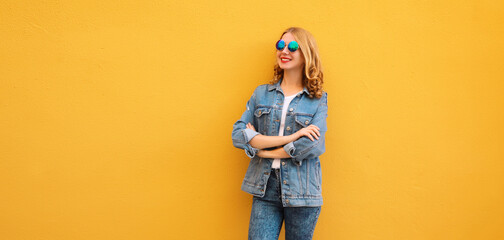 Stylish beautiful happy young woman with crossed arms in denim jacket posing on yellow background
