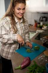 Happy smiling female with excess weight in stylish shirt standing in kitchen chopping broccoli on cutting board with knife, cooking healthy delicious tasty vegetable stew, dieting to keep fit