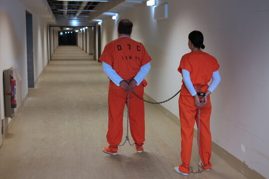weibliche und m&auml;nnlicher Strafgefangene in Handschellen und Fusschellen und orangen Overall mit Sicherungskasten im Kellergang