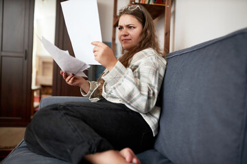 Indoor side view portrait of young frustrated barefoot housewife annoyed with paperwork holding...