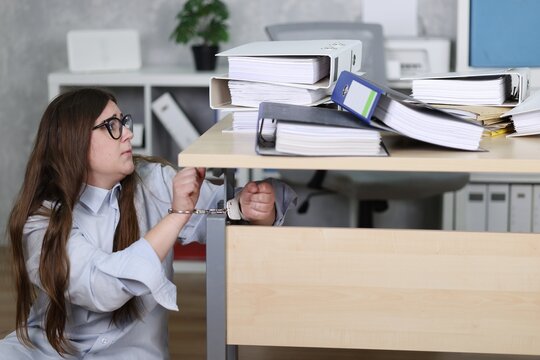 Sekret&auml;rin in Bluse, H&auml;nde in Handschellen, vor viele Aktenordnern im B&uuml;ro, an den Schreibtisch gekettet, auf Boden sitzend