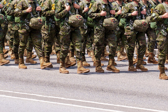 A lines of paratroopers in camouflage uniforms march in formation, carrying parachutes and equipment