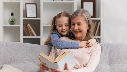 Granddaughter cheerfully covers the eyes of her grandmother who is reading a book sitting on the...