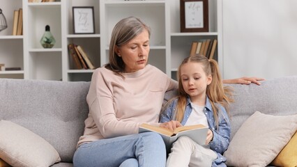 Grandmother and granddaughter are sitting hugging on the sofa and reading a book. A 60s senior grandmother is doing homework with her little granddaughter.