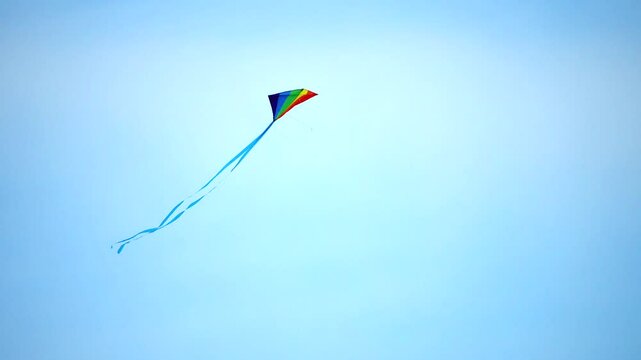A rainbow coloured diamond kite flying in the air, it swoops, twists and turns as the breeze catches it. The long tail trails behind and makes patterns in the sky. Copy space available in the shot
