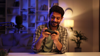 Young confident Arab man playing online video game on his phone, streaming live at home at night, talking into microphone. Gamer lifestyle, E-Sport online gaming technology concept.