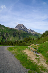 Panorama with a park bench on a hiking trail with a view of the surrounding mountains in the Arlberg area, snow-covered cloudy mountain peaks and green, flower-filled meadows in spring, recreation