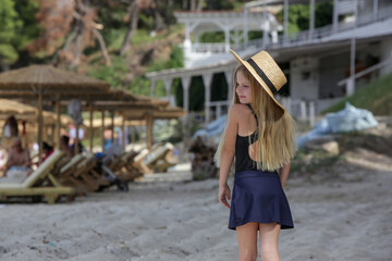 Cute little girl with long blonde hair and straw hat at the resort beach