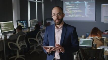 Medium portrait of young Middle Eastern male programming teacher in glasses, jacket using tablet then looking at camera and smiling. School students sitting at computers and learning coding