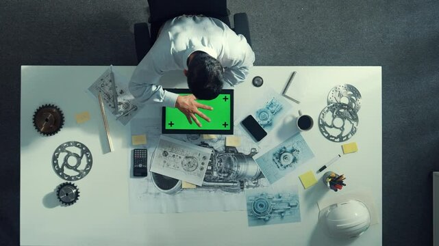 Top view of engineer looking at phone while working on tablet with green screen at meeting table with turbine engine or jet engine sketch design and various metal gear scatter around. Alimentation.