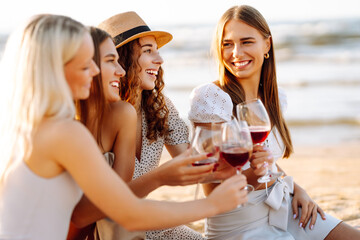Group of young women having fun enjoying a beverage and relaxing on the beach at sunset. Hen party on the beach. Vacation, relax, holiday, picnic.