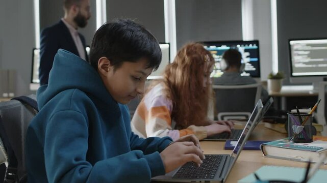Medium portrait of young Indian boy in sweatshirt sitting at programming lesson at school, working on coding project on laptop, looking at camera and smiling, and teacher walking around classroom