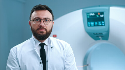 Image of responsible doctor sitting at magnetic resonance scanner. Bearded doctor in glasses. Modern equipped room for MRI scanning. Tomographer in white gown at background of MRI capsule.