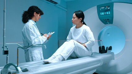 Examination of magnetic resonance imaging at background of modern equipment. Patient sitting at computerized tomography scanner bed, complaining at whole body pain, listening to doctor attentively.
