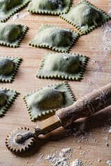 Close-up with handmade spinach ravioli and pastry cutter wheel on floured wooden surface. Vertical composition.