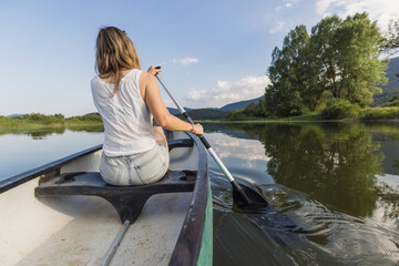 Fototapeta premium Woman paddling the canoe across a beautiful calm mountain lake on a summer sunny day. Canoeing and nature concepts.