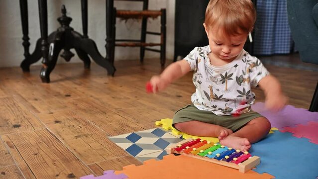 Cute baby boy knocking on colorful iron plates, learning playing educational music toy, Toddler plays with colorful xylophone and educational toys while sitting on a mat indoors. Learning through play