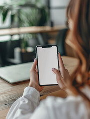 Woman holding a smartphone in her hands with a blank white screen with space for graphics, text or inscriptions on the background of a desk with a laptop
