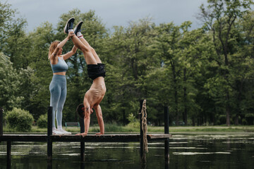 Couple performing fitness exercises on a dock, with a woman assisting a man doing a handstand surrounded by lush greenery