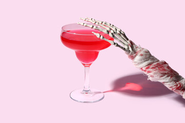 Skeleton hand with glass of tasty cocktail for Halloween celebration on pink background