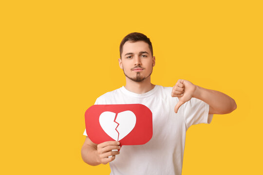 Handsome young displeased man with heart dislike icon showing thumb-down gesture on yellow background