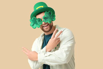 Happy young man with party glasses and leprechaun's hat showing victory gesture on beige...