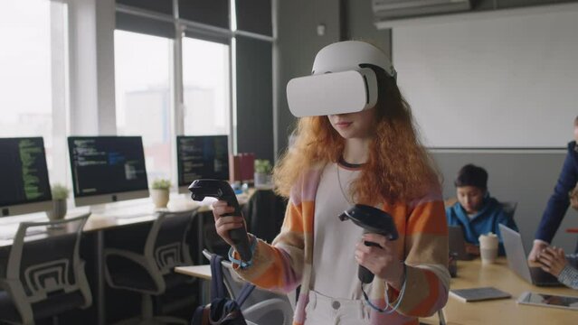Medium shot of teenage Caucasian girl with red hair, wearing VR helmet, exploring metaverse using controllers, looking around, while studying at programming club, teacher helping boy in background