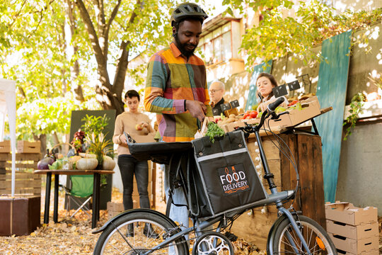 Organic produce delivery services. Picking up food orders at a local farmers market stand and delivering fresh organic vegetables and fruits. On his bicycle, a courier transports farm produce.