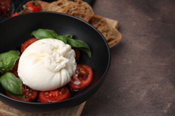 Delicious burrata cheese, tomatoes and basil in bowl on brown table, closeup. Space for text