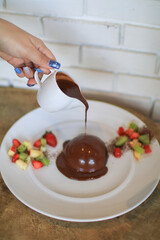 Pouring chocolate sauce over chocolate dome decorated with strawberry and kiwi in white plate. Delicious fresh dessert.