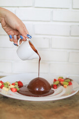 Pouring chocolate sauce over chocolate dome decorated with strawberry and kiwi in white plate. Delicious fresh dessert.