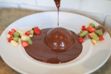 Pouring chocolate sauce over chocolate dome decorated with strawberry and kiwi in white plate. Delicious fresh dessert.