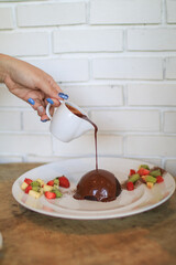Pouring chocolate sauce over chocolate dome decorated with strawberry and kiwi in white plate. Delicious fresh dessert.