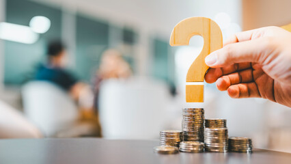 A businessman's hand holds a question mark on a pile of money placed on a table with a blurred...