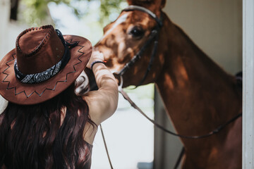 A woman wearing a cowboy hat prepares her horse for a ride outdoors. The moment captures a close...