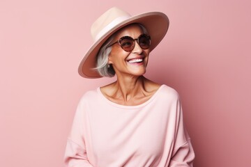 Portrait of a beautiful senior woman in hat and sunglasses smiling against pink background