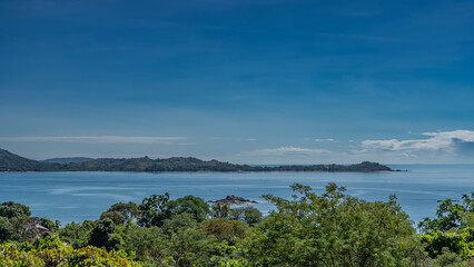 A tiny rocky island is visible in the bay. The blue ocean is calm. Hills in the distance against the background of sky and clouds. There is green vegetation in the foreground. Madagascar. Nosy Iranja