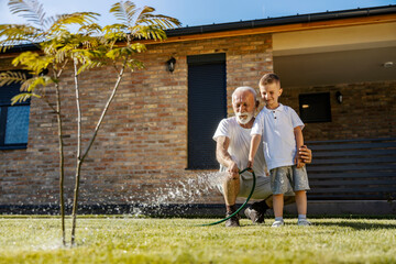 Happy grandfather is hugging his grandson in backyard and watering lawn and tree together.