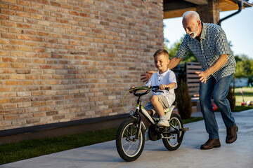 Happy child is learning how to ride a bicycle and his granddad is helping and running in backyard.