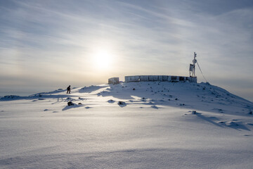 Ruins of an abandoned structure on the top of a mountain. In the distance, a woman climbs a snow-covered mountainside. There is a solar halo in the sky. Cold weather. Magadan region, Russian Far East.