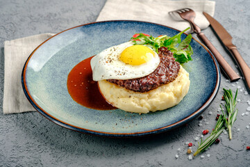 Juicy beef patty with fried egg and mashed potatoes with gravy on a blue plate decorated with rosemary