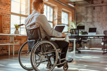 man in a wheelchair is sitting at a desk with a laptop. The scene is set in a large office with a potted plant in the background. The man is working on his laptop, possibly for a job