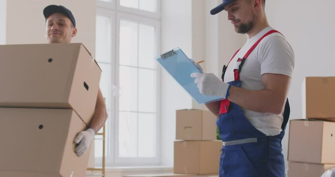 Young man worker from moving service standing with clipboard making notes for movers working in client house carrying cardboard boxes. Move, moving day, delivering and relocation concept. 4k video.