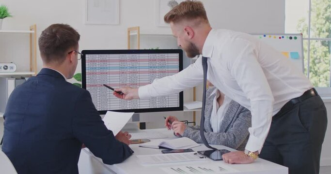 Team of business people meeting by a table in a modern office interior, discussing work matters, using a desktop computer and a tablet, looking at some financial data sheets