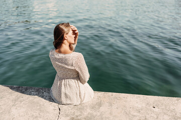 A woman is sitting on a ledge by a body of water. She is wearing a white dress and she is looking out at the water. The scene is peaceful and serene, with the woman taking a moment to enjoy the view.