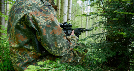 Hunter with rifle wearing camouflage suit sitting in ambush. Camouflaged sniper in the forest in...