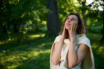 Fototapeta premium Young pretty woman in white modern casual clothes walking in the park, wandering around in nature and relaxing. Cute girl in white hoodie.