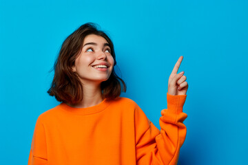 A young woman in an orange sweater pointing upwards against a blue background, with copy space for adding text or messages