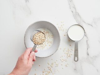 Hand pouring oat flakes over bowl of milk preparing porridge for breakfast. On the table half a...